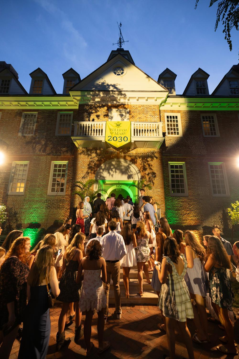 New students walk up the stairs into the Wren building at twilight with a "2030" banner hanging on the balcony