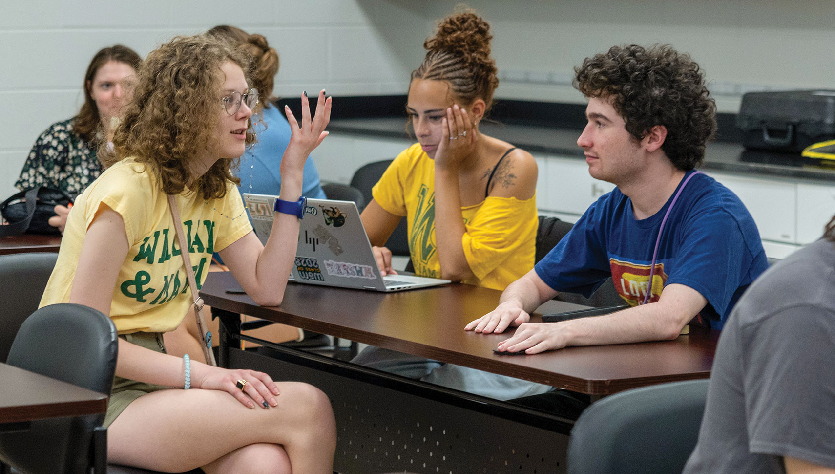 Three students collaborate around a classroom table, discussing ideas beside an open laptop