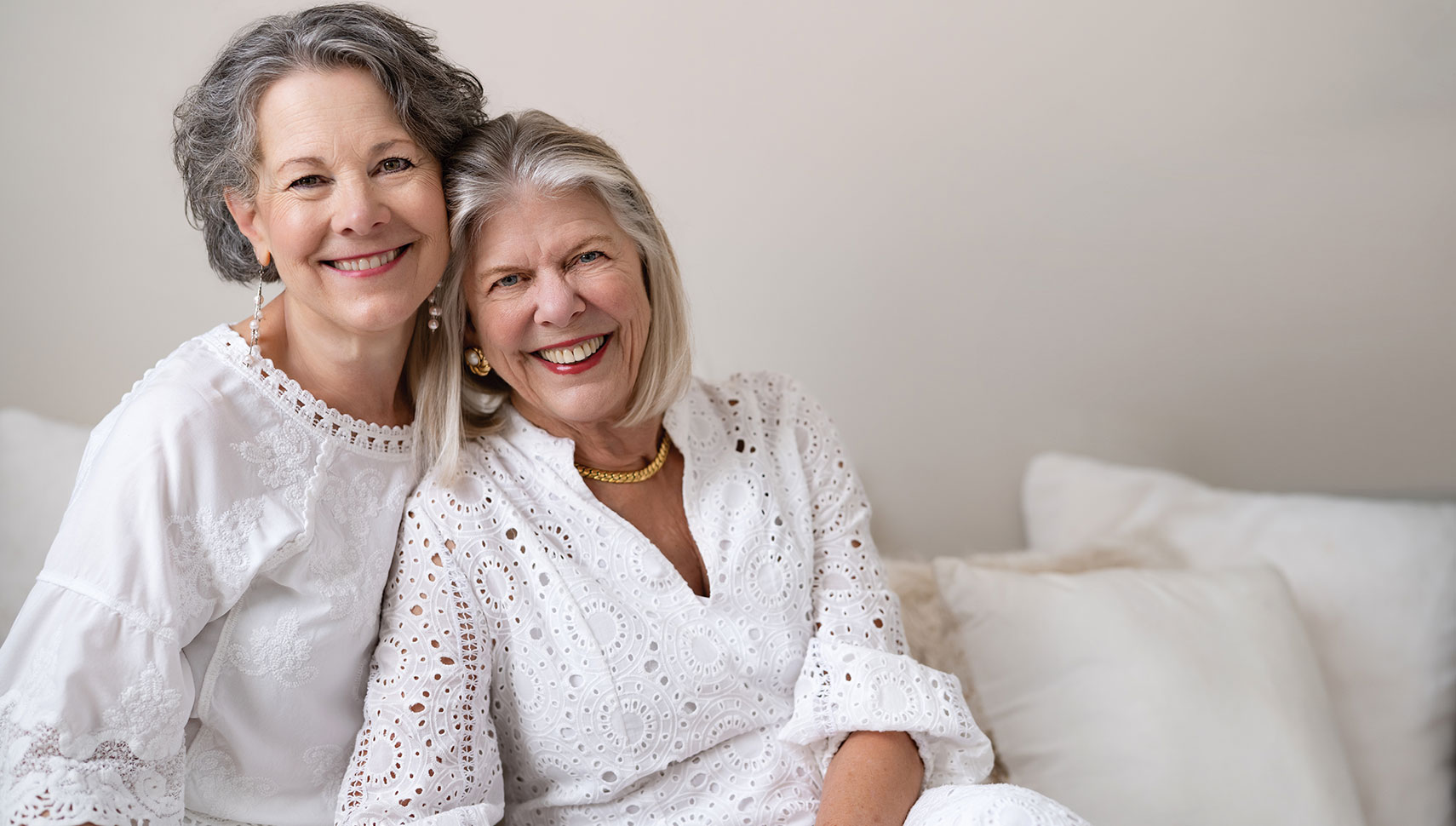 Mary Beth Morrill Gibson and René Roark Bowditch sit close together on a couch, leaning toward the camera in a bright, warm portrait.