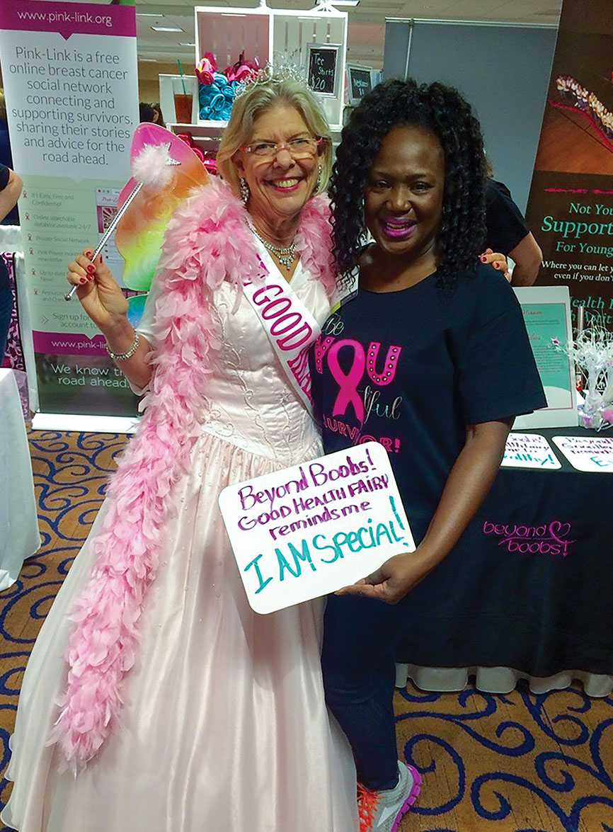 Two smiling women pose at a breast cancer support booth; René Roark Bowditch wears a white gown, pink feather boa and sash, holding an “ Beyond boobs, Good Health Fairy reminds me I am special” sign.