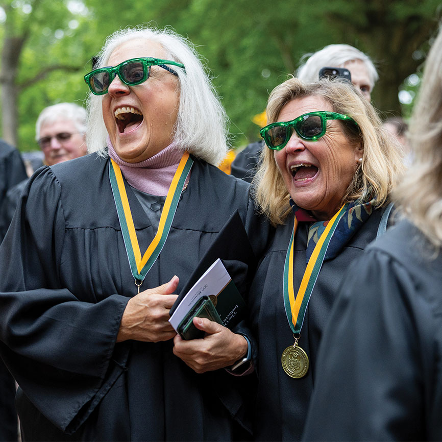 two women in graduation gowns, wearing an olde guarde medallion and light up sunglasses