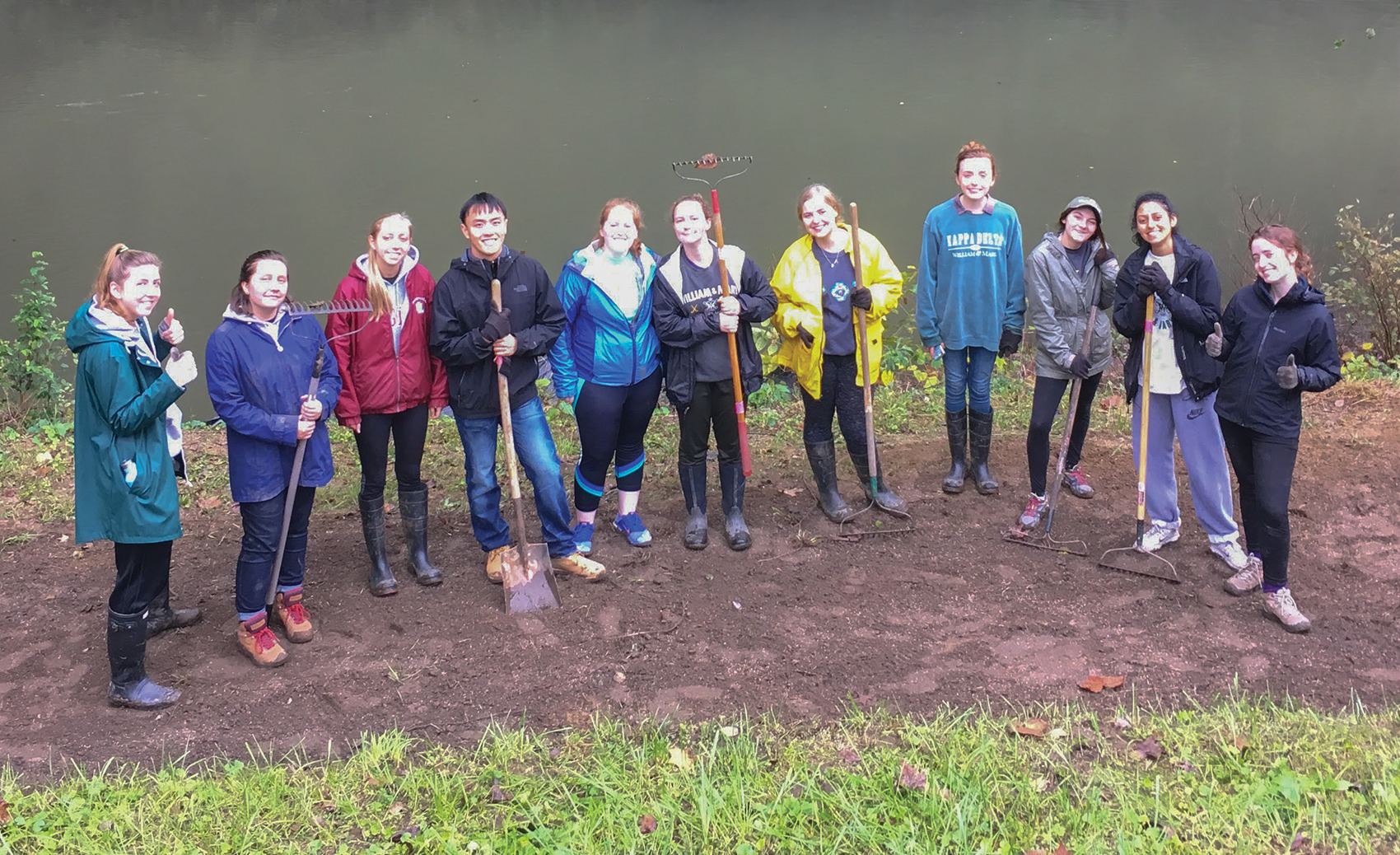 11 students in raincoats stand in a line with rakes on a dirt river bank