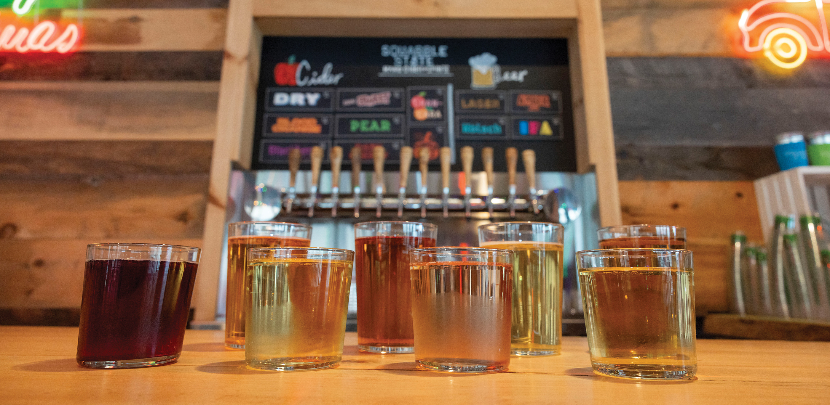 An array of varying color of ciders in glasses line a bar top