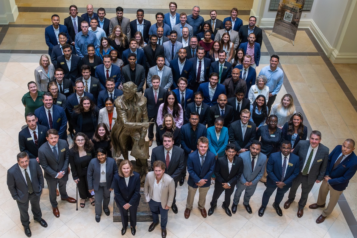 Large group of business school staff and faculty posing around a bronze statue in a bright atrium with Jill Ellis and Todd Howard  