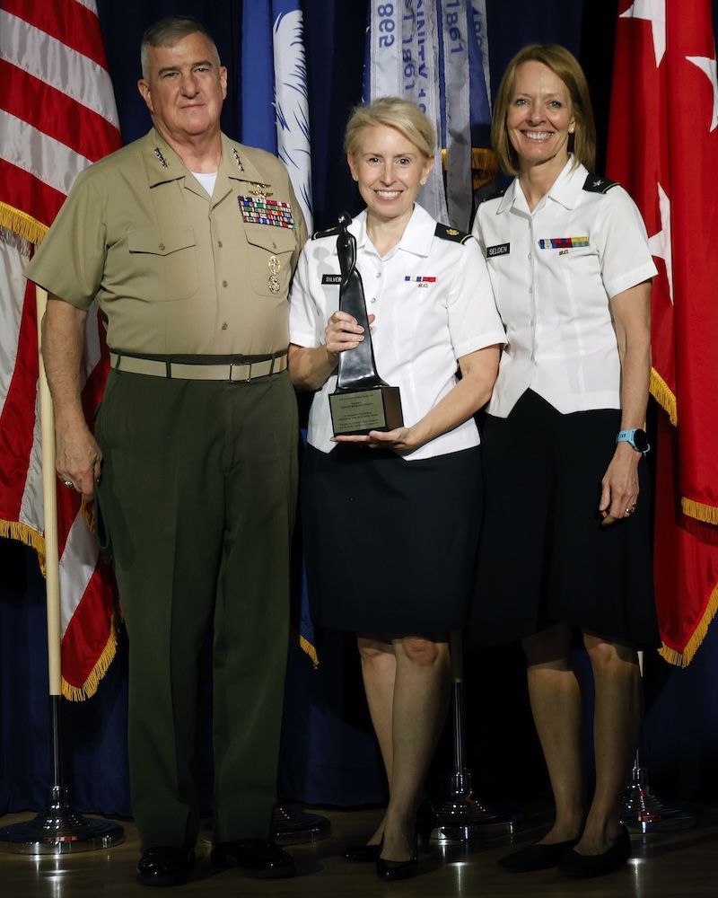 Silverman holding his award at the ceremony, standing between two men in dress uniform and business attire.