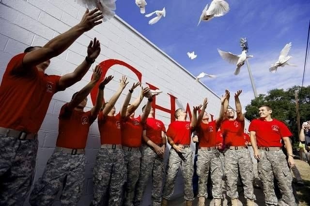A group of people in red shirts and camouflage pants release white doves beside a white wall