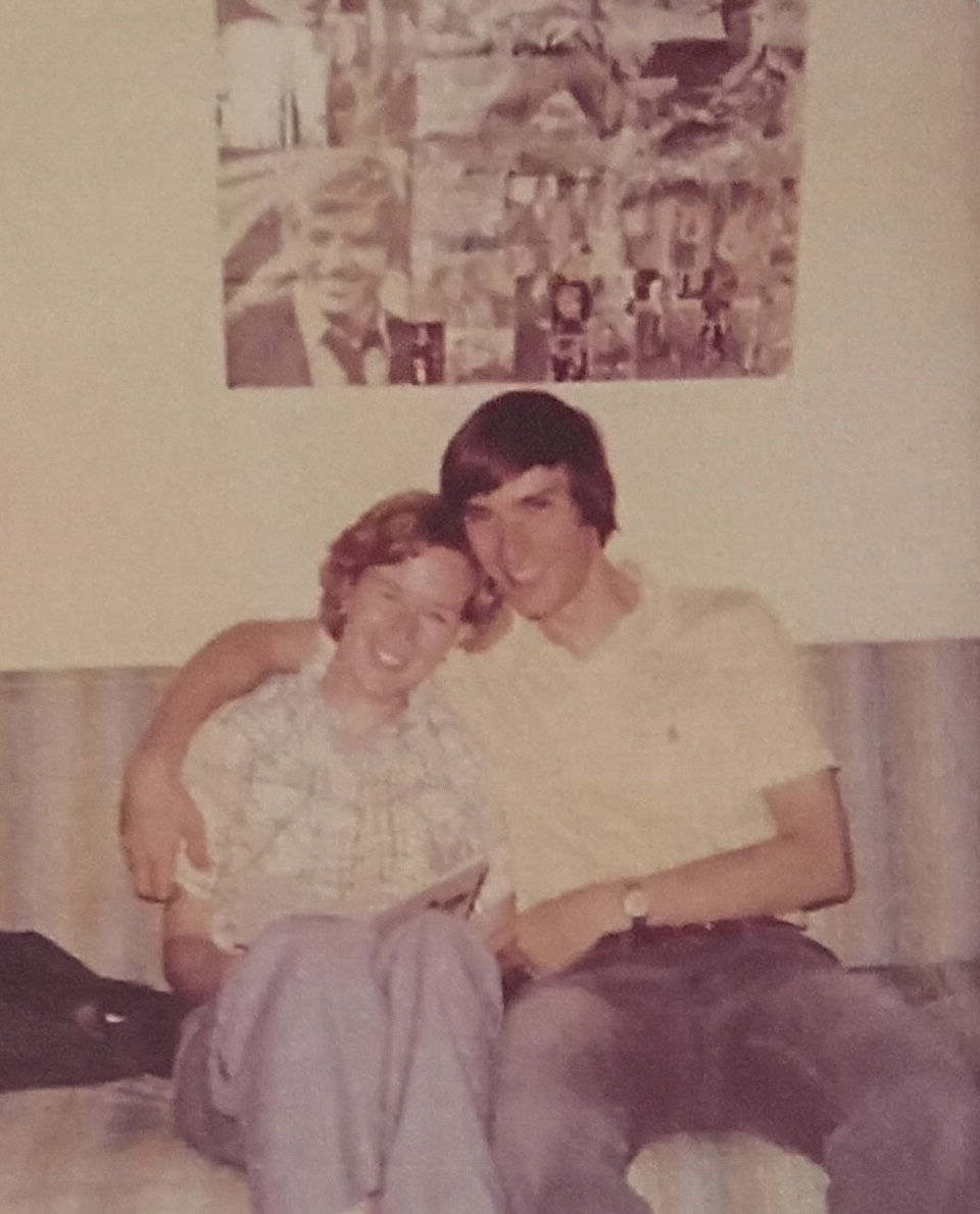A 1970s photo of a couple sitting in front of a collage on a wall of black and white photos of Robert Redform